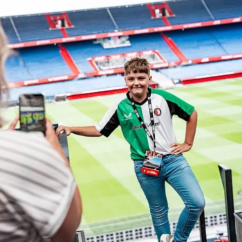 A boy wearing a green and white jersey poses for a photo in a stadium, with a person capturing the moment on a phone.