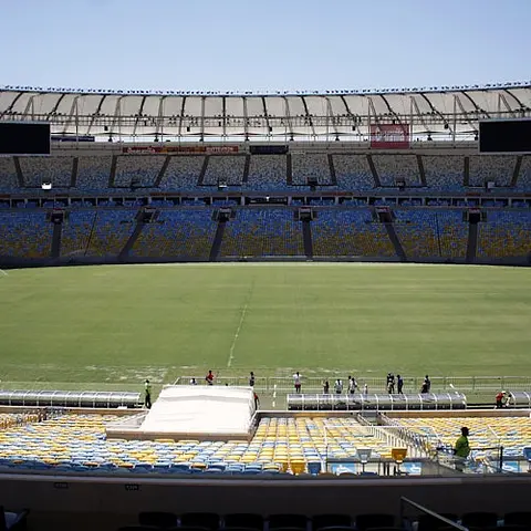 Un stade vide avec des sièges multicolores, un terrain vert et quelques personnes debout près de la ligne de touche sous un ciel clair.