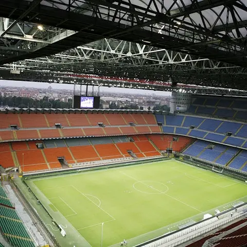 Stade de football vide avec un terrain vert, des sections de sièges rouges et bleus, et un grand écran au-dessus d'une extrémité. Le ciel est partiellement visible.