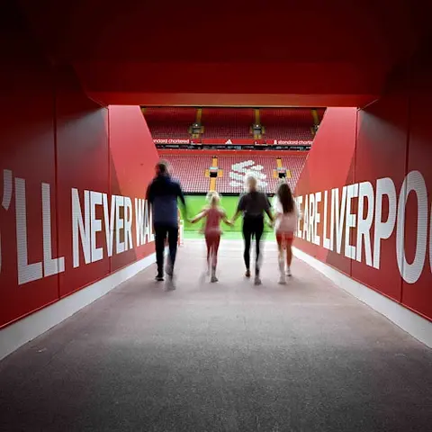Quatre personnes se tiennent par la main et traversent un tunnel rouge en direction d'un terrain de football. Le texte sur les murs dit "You'll Never Walk Alone" (Vous ne marcherez jamais seul).