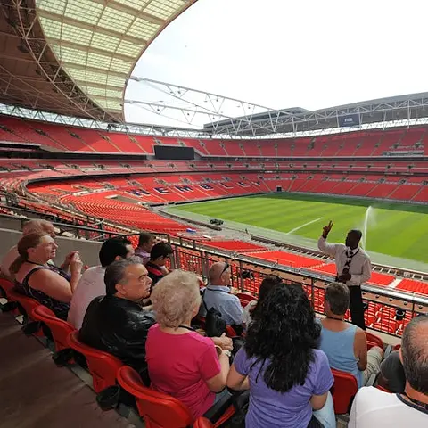 People seated in a stadium tour, listening to a guide, overlooking an empty football field with red seats and overhead roof.