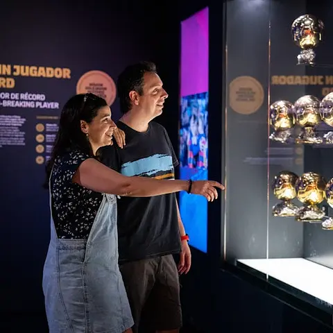 Two people smile and point at a display of multiple golden soccer ball trophies inside a dimly lit exhibit.