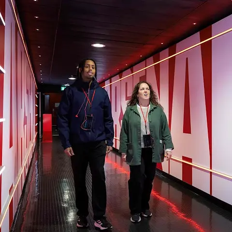 Two people walking through a well-lit corridor with red and white walls featuring large text, wearing casual clothing.