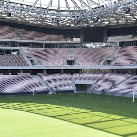 Leeres Stadion mit abgestuften Sitzplätzen und einem gitterartigen Dach, mit Blick auf einen gut gepflegten Rasenplatz. Keine Menschen anwesend.