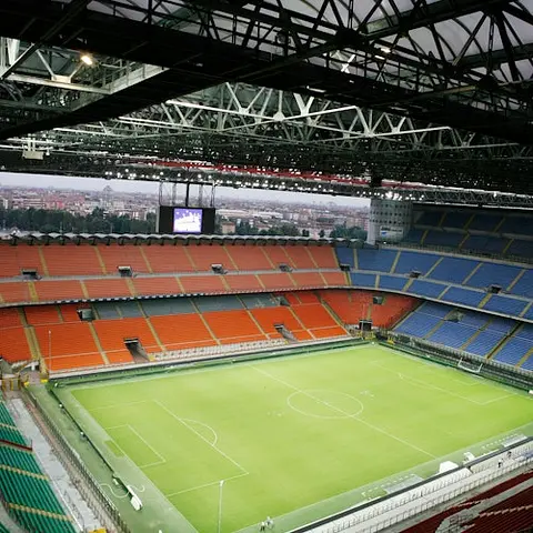 Empty football stadium with a green field, red and blue seating sections, and a large screen above one end. Sky partially visible.
