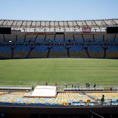 Estadio vacío con asientos multicolores, campo verde y unas pocas personas de pie cerca de la línea de banda bajo un cielo despejado.
