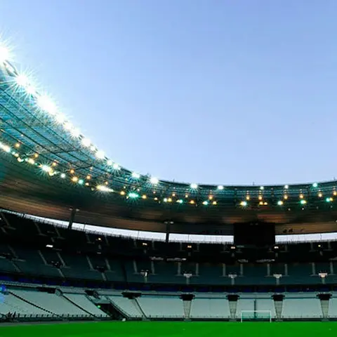 Ein leeres, großes Stadion mit abgestuften Sitzreihen und hellen Lichtern, über dem ein grünes Feld und ein klarer Himmel zu sehen sind.
