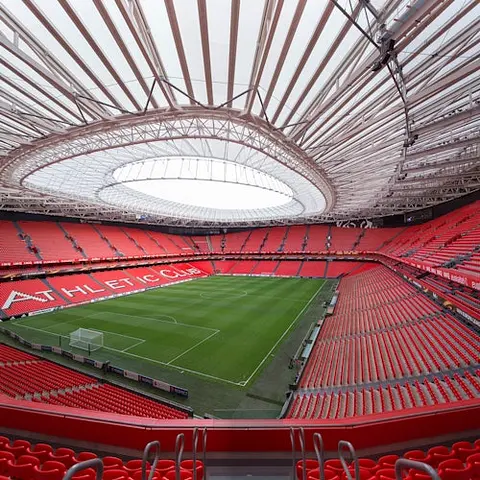 Leeres Fußballstadion mit roten Sitzen, großem Dach und der Aufschrift "ATHLETIC CLUB" auf der Tribüne. Grünes Spielfeld in der Mitte.