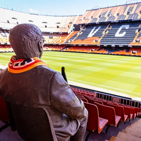 Una estatua sentada en las gradas de un estadio de fútbol vacío, llevando una bufanda con los colores rojo, amarillo y negro.